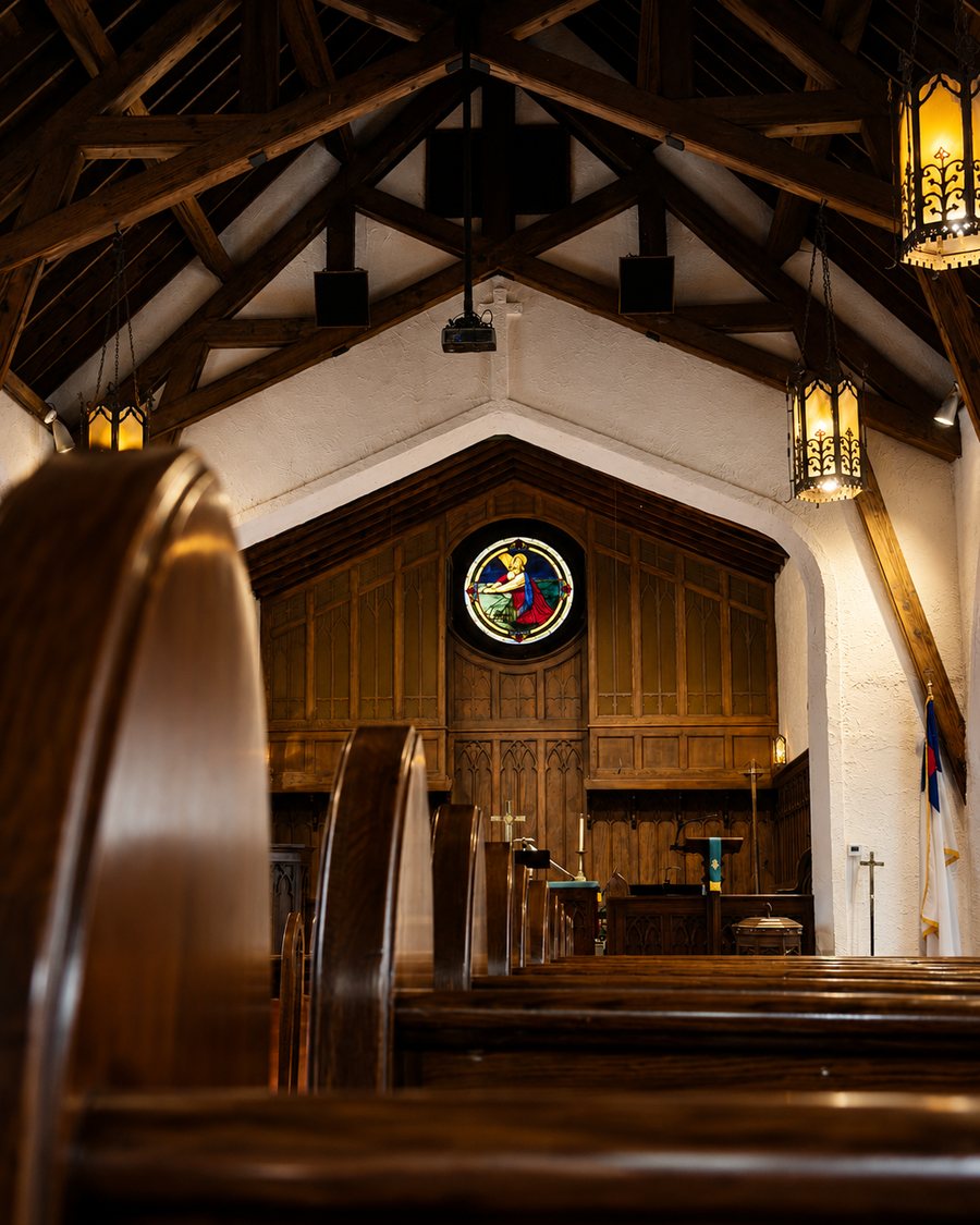 The warmly lit sanctuary of Lambuth Memorial United Methodist Church — exposed wood trusses, glowing lanterns, and a stained-glass rose window above the chancel.